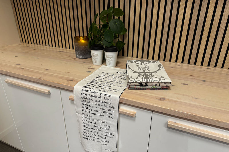 Ljust nordiskt kök med träbord, vikta kökshanddukar i linne och bomull i mjuka färger. En handduk hänger bredvid koppar med renmotiv och en liten vas med växt. Scenen fångar en varm, skandinavisk känsla av enkelhet och naturlig design.